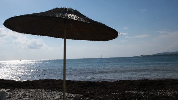A straw umbrella on a beautiful pebble beach. Summer straw umbrella and the reflection of the sun in rippled water surface, summer holiday concept.