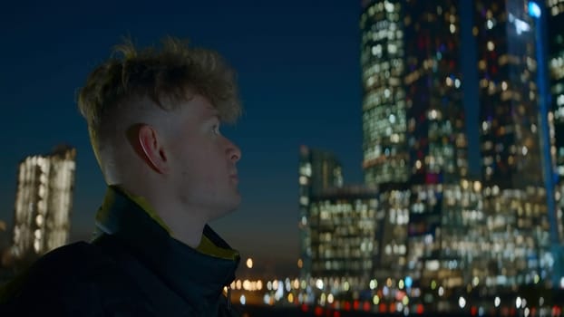 Man looks at modern glowing skyscrapers at night. Action. Young man on background of bright glowing skyscrapers at night. Man looks at skyscrapers glowing at night in city.