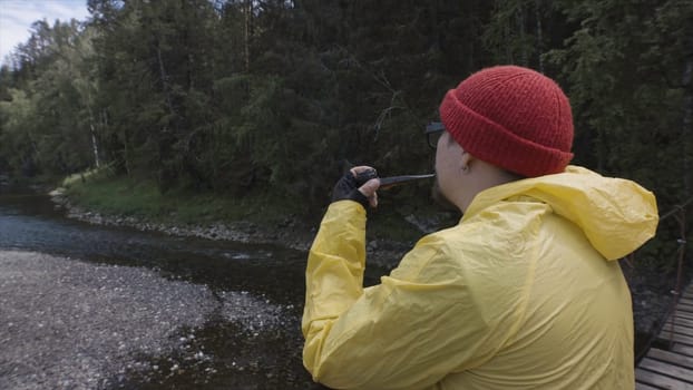 Side view of the bearded man in warm red hat smoking pipe in the wild forest in the autumn. Man in yellow jacket standing on hanging bridge above the river.