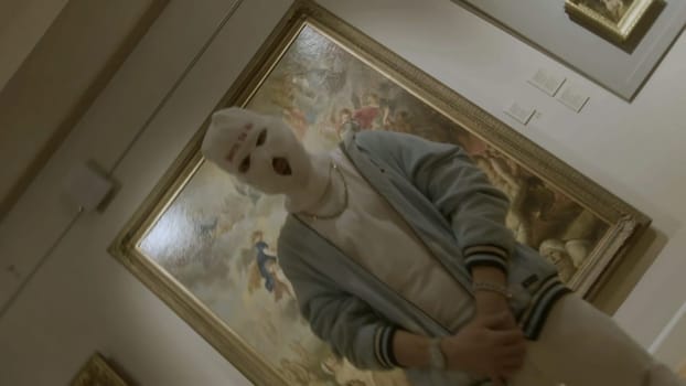 A man in balaclava at the art exhibition near paintings. Footage. Concept of vandalism, man at art museum
