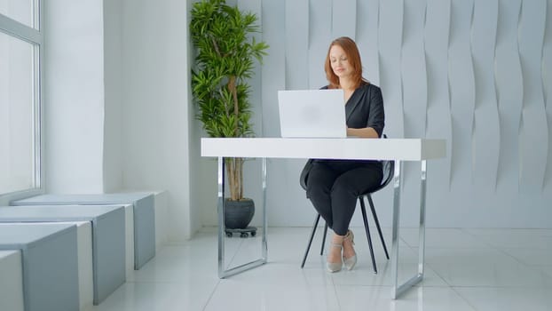Working woman with red hair. Action.A woman in a black suit in a cozy room who is sitting at a computer desk in a laptop working. High quality 4k footage