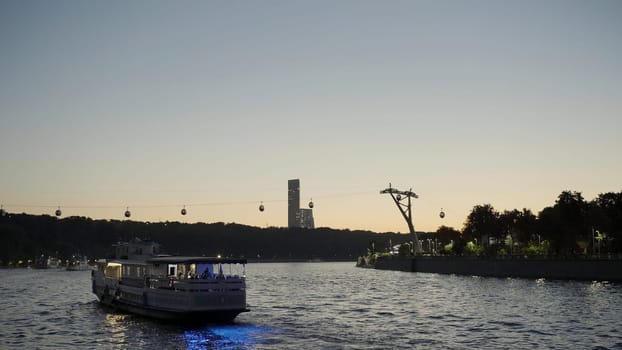 Sunset landscape with a touristic yacht sailing along embankment. Action. Cable car moving above the river
