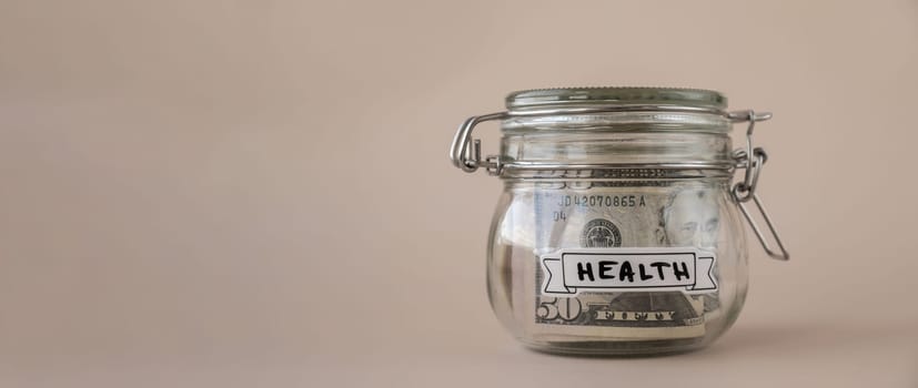 Saving Money In Glass Jar filled with Dollars banknotes. HEALTH transcription in front of jar. Managing personal finances extra income for future insecurity. Beige background