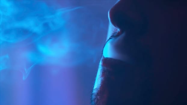 The mouth of a young man smoking the traditional hookah in the dark room, bad habits concept. Close up of male face detail, exhaling smoke.