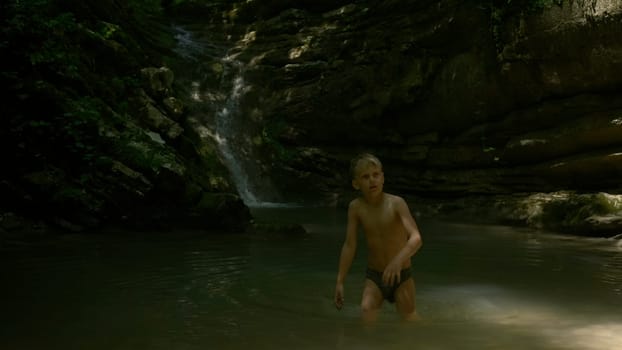 Boy kid having a great time in the cold mountain stream. Creative. Beautiful waterfall and big cliffs