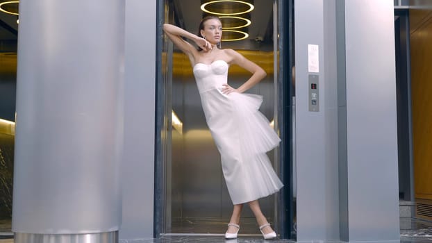Woman in white dress poses on background of lift. Action. Elegant young woman poses inside modern building. Beautiful woman in elegant white dress posing.