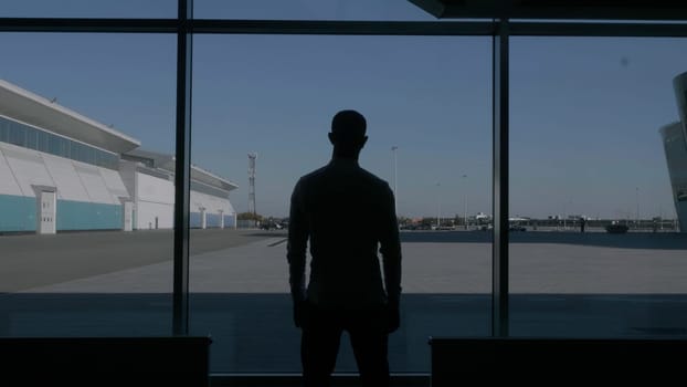 Back view of a confident successful male silhouette in front of a large window. HDR. Thoughtful businessman watching the summer blue sky and a parking space