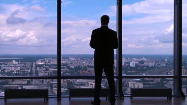 Businessman in suit standing next to a big window from an office building and checking his watch back view. Businessman standing at window looking on his wrist watch.