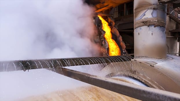 Red hot melted metal flowing inside the chute. Industrial background at the factory hot shop with flowing hot steel near rotating mill being cooled by water.