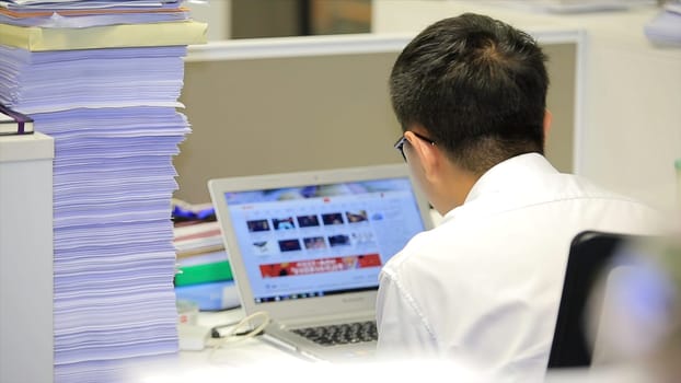 Asian man working on a laptop. Young pensive Asian man working on laptop at office, business or technology concept. View from back.