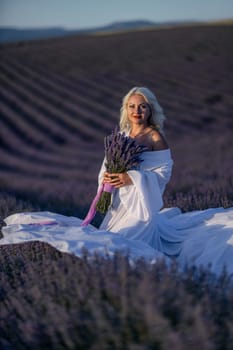 Blonde woman poses in lavender field at sunset. Happy woman in white dress holds lavender bouquet. Aromatherapy concept, lavender oil, photo session in lavender.