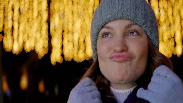 The girl in the gray hat. Action. A woman who stands on a dark street with bright yellow garlands burning behind her and who makes a face at the camera while adjusting her neck. High quality 4k footage