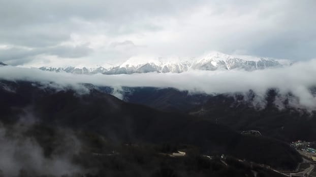 Aerial view of the stunning winter landscape of the snowy slopes and Alps hills among the lush white clouds. Flying over the green forest and snowy peaks in the distance.