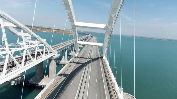 Huge bridge. Drone view. Action. A long bridge over the ocean on which cars can move freely, the bridge is located next to the green mountains.
