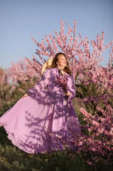 Woman blooming peach orchard. Against the backdrop of a picturesque peach orchard, a woman in a long pink dress and hat enjoys a peaceful walk in the park, surrounded by the beauty of nature