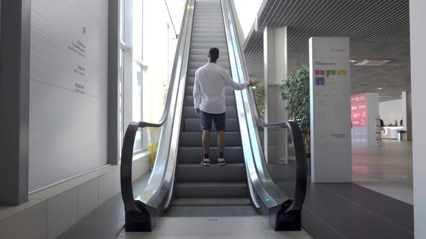 Young man climbs escalator in business building. HDR. Rear view of man climbing on escalator. Man climbs an escalator in bright building with large windows.
