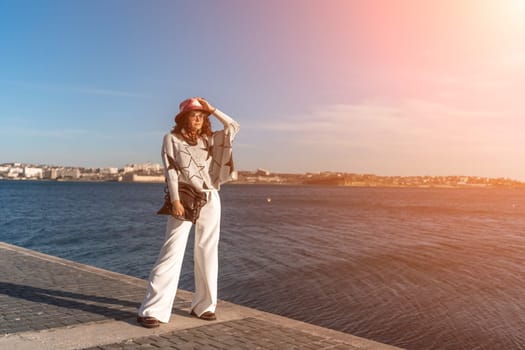 Stylish seashore woman. Fashionable woman in a hat, white trousers and a light sweater with a black pattern on the background of the sea