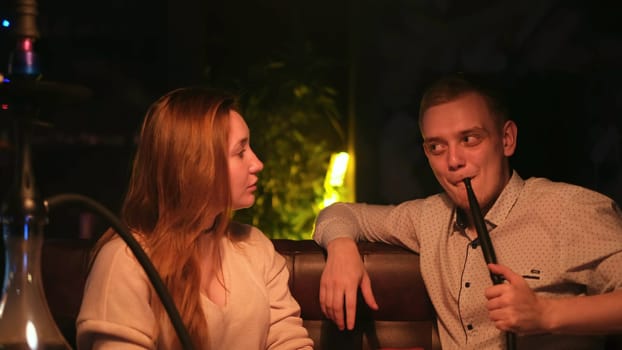 Young loving couple smoking a hookah while sitting in a cafe by the green neon lamp. Young husband and wife enjoying shisha and chatting.