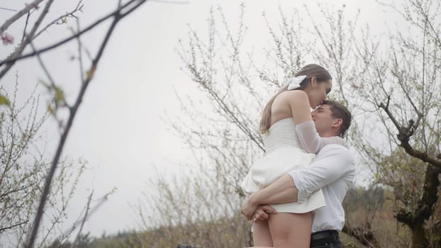 Beautiful couple embraces in spring garden. Action. Couple of newlyweds enjoy first leaves and flowers in garden in spring. Couple embraces in blooming garden in early spring.