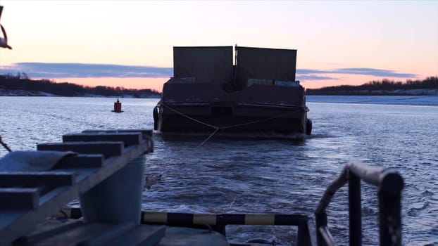 Barge in tow of fishing boat. Clip. Boat carries barge with cargo in cloudy twilight. Boat and barge float on river on winter morning.