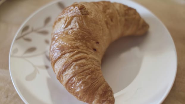 Top view of tasty fresh croissant lying on the white glass plate, french breakfast concept. Close up of beautiful pastry product on the wooden surface.