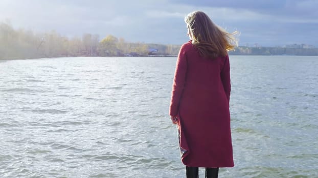 Young girl with her hair stands near the water in the cold season. Back view. Young woman standing near a lake.