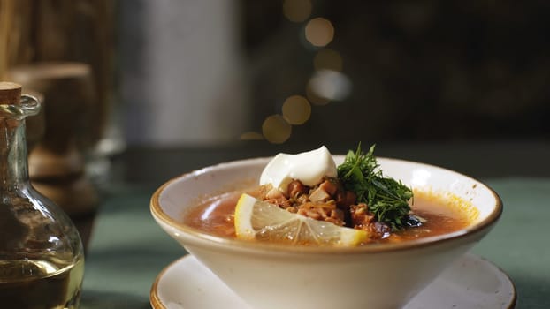 Close-up of hand putting the plate with Russian traditional soup solyanka on the table. Russian solyanka soup with meat, sausages and olives