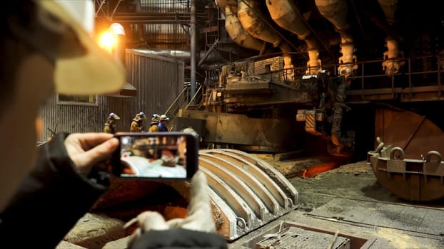 Rear view of the engineer in hardhat is using a phone to shoot a video inside industry factory. Metallurgical plant and professional equipment.