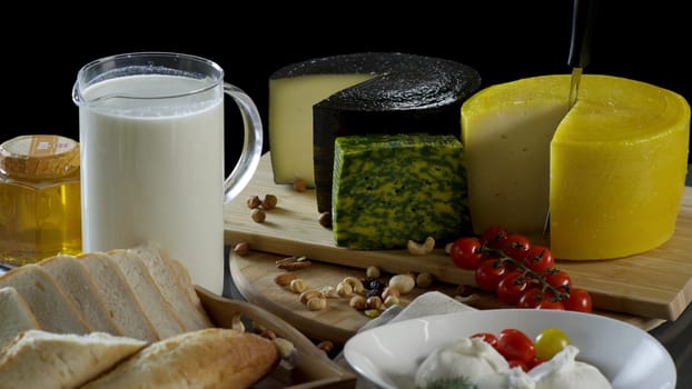 Still life in the old style with a jug and a mug of milk, a heads of cheese, toast, honey and eggs on black background. Cheese camembert, blue cheese, parmesan, toasts, honey and walnut on a dark background.