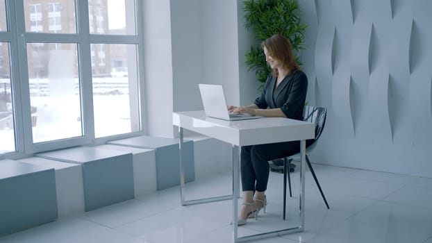 Beautiful woman is sitting at desk in office. Action. Businesswoman is typing on laptop in stylish modern office. Woman works in modern office.