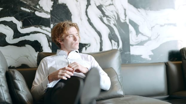 Handsome man in suit sits down on sofa. Action. Handsome man in white shirt confidently sits down on sofa. Man sits down on sofa with expression of confidence and thoughtfulness.