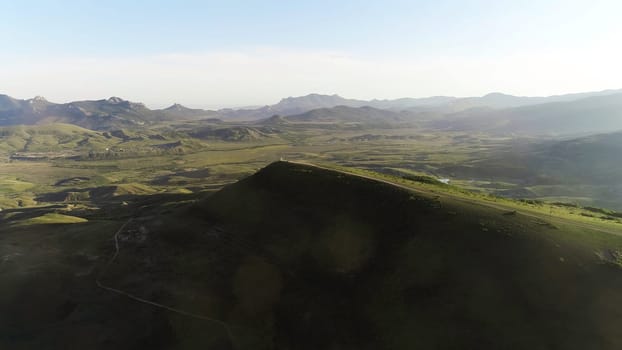 Aerial view of country road on the top of high green hill with meadows and mountain chain on the background against blue sky. Beautiful view from above