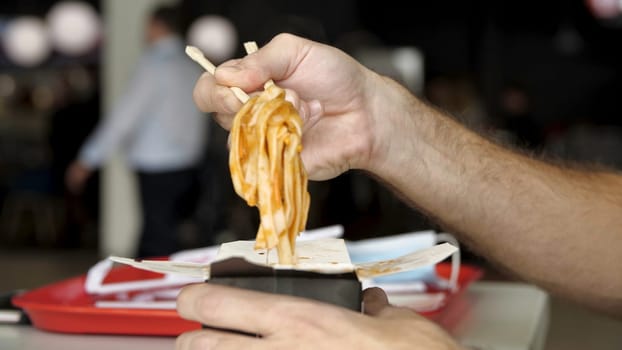 Close up of male hands with wooden chinese chopsticks eating delicious asian wok noodles. Side view of eating chinese pasta in a public place.