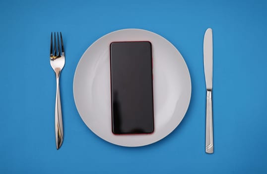 Mobile phone lying in white plate near fork and knife. Phone addiction concept