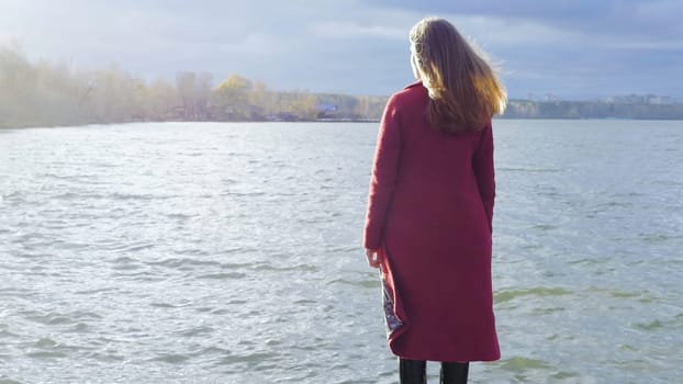 Young girl with her hair stands near the water in the cold season. Back view. Young woman standing near a lake.