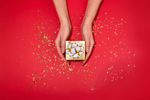Female hands holding small gold box with a white bow gift hidden inside on red maroon background with scattering of gold sparkles and glitter top view Christmas New Year Valentine's day birthday