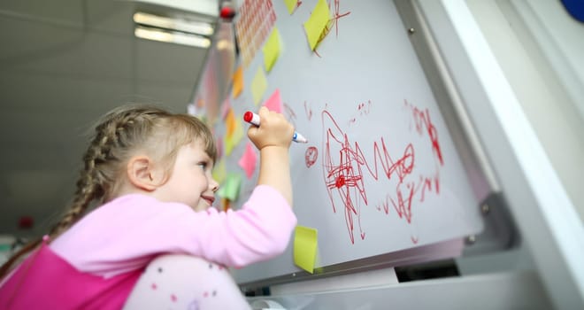 Portrait of joyful preschooler holding big red marker and drawing something abstract. Happy girl having fun and standing in modern classroom in trendy pink sweater. Creative childhood concept