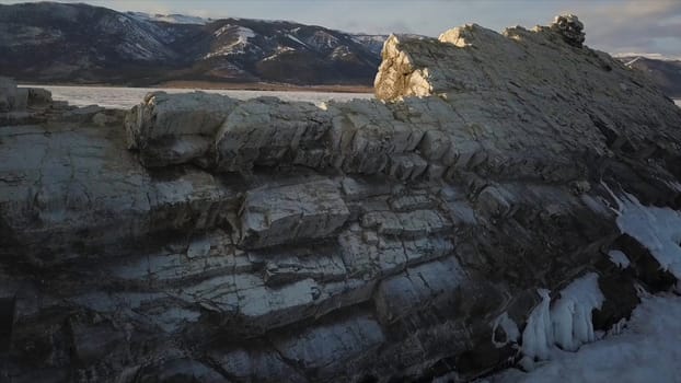Aerial view of the cliff with steep slopes on the background of forested mountains and frozen pond. Mountains with peaks partially covered with snow by the beautiful frozen Baikal Lake under blue sky.