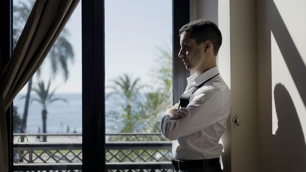 Side view of a man in white suit standing by the window. Action. Sea and palm trees behind the window