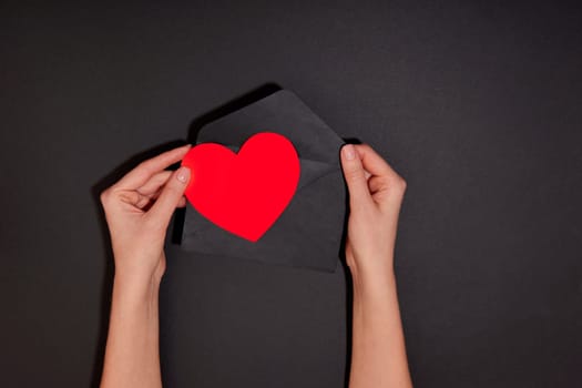 Female hands takes out and receives or sends a red card in the shape of a heart from an envelope on a black background, top view copy space