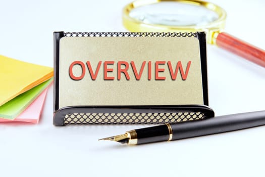 OVERVIEW text on a gold plastic card with convex text next to office supplies on a white background