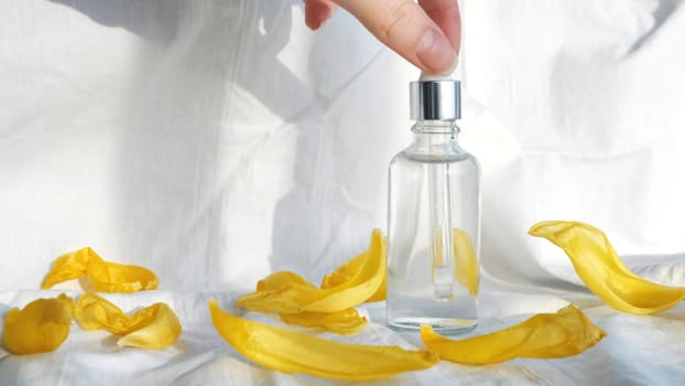 A serum and dropper on a white textile background, natural cosmetics. Close up of yellow flower petals and tender female hands touching small glass bottle with oil inside.