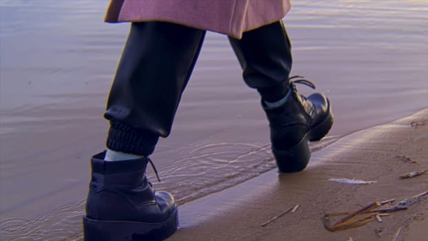 Close up of female leather black boots walking on a sandy shore and the water edge. Details of a young woman walking by the river.
