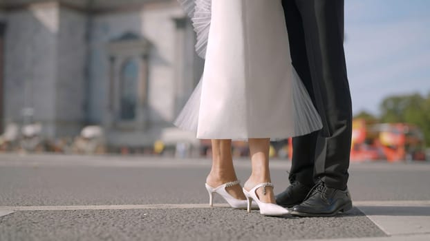 Legs of stylish bride and groom on the background of city summer street with driving cars. Action. Man in black suit and a woman in white dress