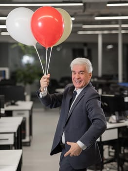 Portrait of a cheerful mature business man holding balloons in the office. Vertical photo