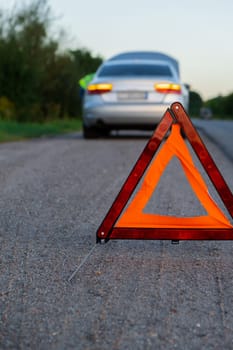 Unrecognizable sad driver in reflective vest. Male driver standing near broken car with open up hood. Red triangle to warn other road users of car breakdown or engine failure stop at countryside highway. Emergency traffic situation