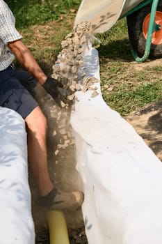 A man pours rubble crushed stone from a wheelbarrow into a trench. Drainage works for drainage of ground water around the field.