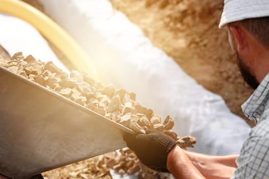 Drainage works for drainage of ground water around the field. A man pours rubble crushed stone from a wheelbarrow into a trench close up.