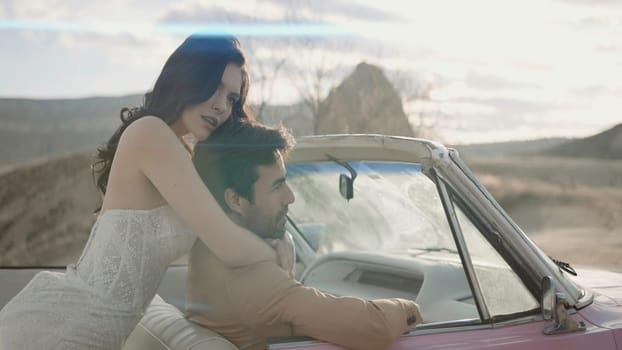 An interesting beautiful photo shoot of the newlyweds. Action. The bride of pretty appearance in a narrow beautiful white long dress with her cute groom in a brown suit are photographed posing next to a pink car hugging and looking at each other