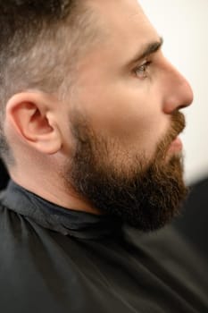 A young man with a beard in a black cape sits on a chair in a barbershop. The client is waiting for a haircut and shaving of the beard.